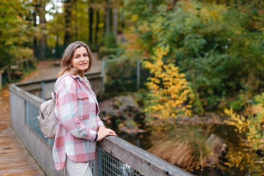 Portrait of cute young woman in casual wear in autumn, standing on bridge against background of an autumn Park and river. Pretty female walking in Park in golden fall. Copy space. smiling girl in the park standing on wooden bridge and looking at the camera in autumn season