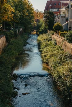 Beautiful small river with clean and clear water front of colorful autumn trees and small old town on the hill agaist nice blue and clouds sky during autumn in Europe.