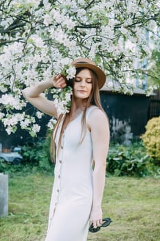 Beautiful young girl in white dress and hat in blooming Apple orchard. Blooming Apple trees with white flowers