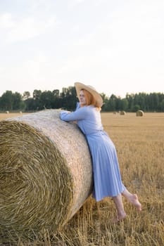 A red-haired woman in a hat and a blue dress walks in a field with haystacks