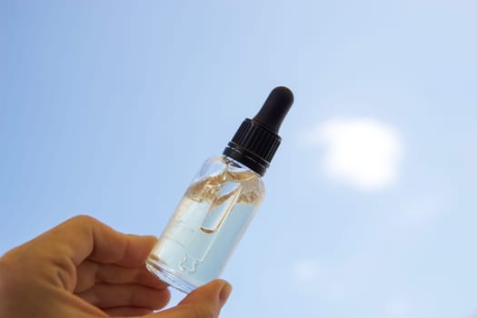 A bottle of cosmetic serum or gel in a woman's hand against a background of blue sky, clouds and sun. The concept of summer holidays and the use of sunscreens.