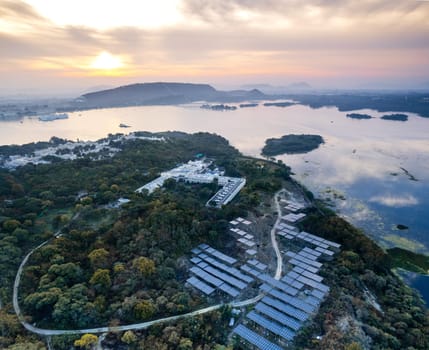 Aerial drone shot with solar panels placed in middle of trees on edge of lake reflecting sunrise colors and aravalli hills showing grene ecological renewable energy production Udaipur India