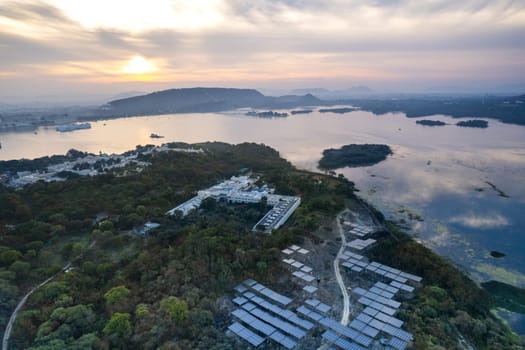 Aerial drone shot with solar panels placed in middle of trees on edge of lake reflecting sunrise colors and aravalli hills showing grene ecological renewable energy production Udaipur India