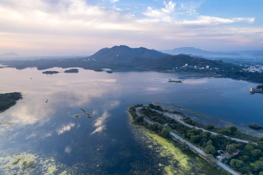 aerial drone shot at dawn dusk with road loop extending into fateh sagar lake with aravalli hills in distance hidden in fog showing cityscape of Udaipur Rajasthan India