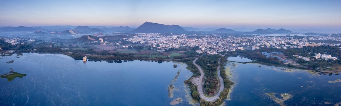 Panoramic aerial drone shot at dawn dusk with road loop extending into fateh sagar lake with aravalli hills in distance hidden in fog showing cityscape of Udaipur Rajasthan India