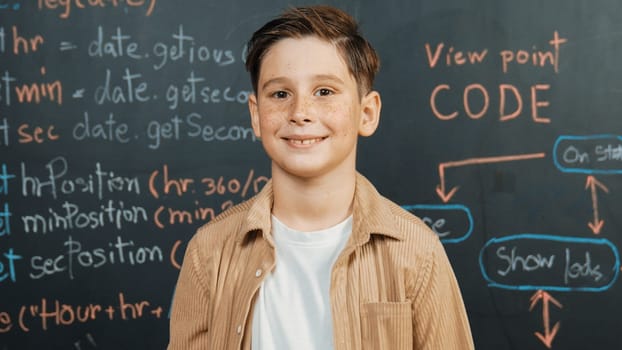 Panorama shot of happy caucasian boy smiling and standing at blackboard with engineering code or prompt written. Smart child looking at camera and greeting while study in STEM classroom. Erudition.