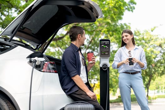 Lovely young couple drinking coffee while recharging battery for electric car during road trip travel EV car in natural forest or national park. Eco friendly travel during vacation and holiday. Exalt