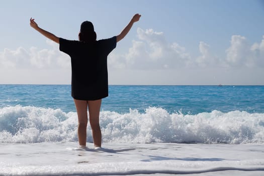 teenage girl in a black T-shirt stands in the sea with her arms outstretched high, rear view.