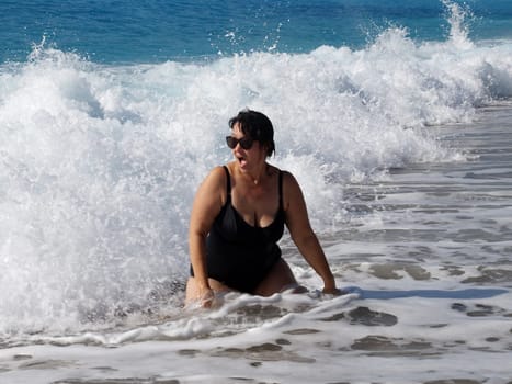 curvy woman in a black swimsuit and sunglasses sits in the sea under the lapping waves.