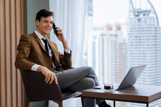 Closeup of handsome businessman making phone call with manager while sitting near window with skyscraper view. Executive manager talking working by using phone and laptop. Look aside. Ornamented.