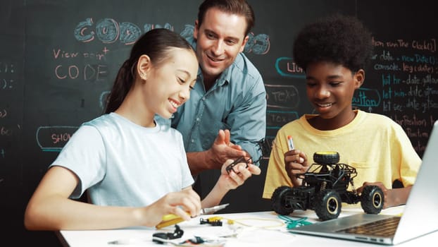 Caucasian teacher looking at laptop screen while explain car model system at blackboard with coding or programing code written. Happy diverse student learning about robotic construction. Edification.