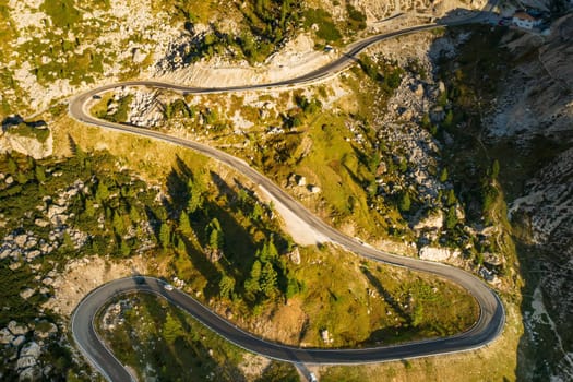 Curvy road winds in highland among forests and hills at sunset. Cars drive on serpentine highway in scenic mountains in sunny evening aerial view