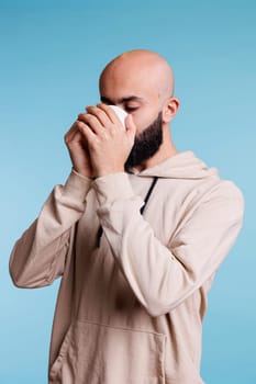 Arab man drinking coffee while holding white mug with two hands. Young adult bald beared person wearing casual beige hoodie while enjoying hot tea from cup, sipping beverage