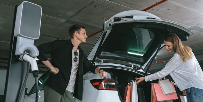 Young couple travel with EV electric car to shopping center parking lot charging in downtown city showing urban sustainability lifestyle by green clean rechargeable energy of electric vehicle innards