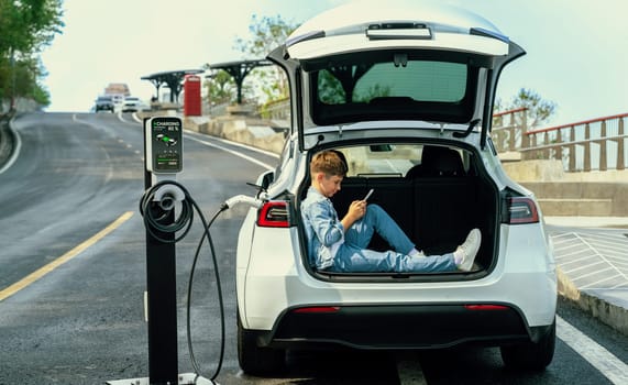 Little boy sitting on car trunk, using smartphone while recharging eco-friendly car from EV charging station. EV car road trip travel as alternative vehicle using sustainable energy concept. Perpetual