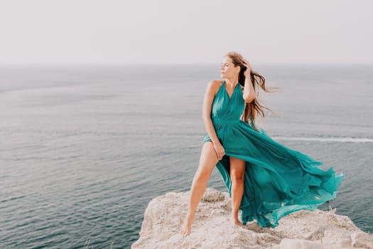 Side view a Young beautiful sensual woman in a mint long dress posing on a volcanic rock high above the sea during sunset. Girl on the nature on overcast sky background. Fashion photo