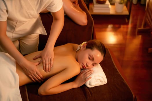 Caucasian couple customer enjoying relaxing anti-stress spa massage and pampering with beauty skin recreation leisure in warm candle lighting ambient salon spa at luxury resort or hotel. Quiescent