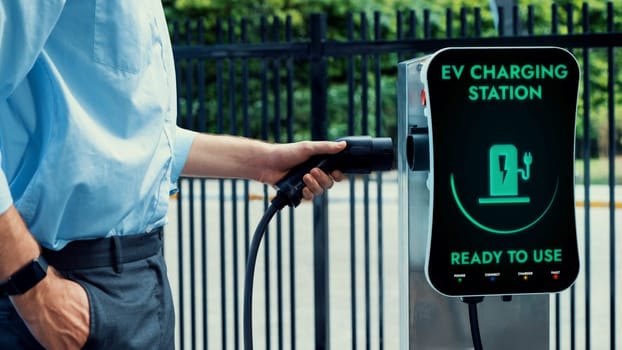 Businessman pull and hold EV charger plug form electric car charging station at city car park area background. Futuristic clean sustainable energy and EV car technological advancement.Peruse