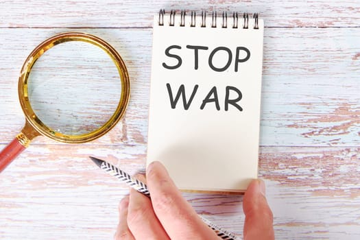 STOP WAR written on a blank sheet of a notebook which is touched by a man's hand with a pencil and a magnifying glass is lying next to it