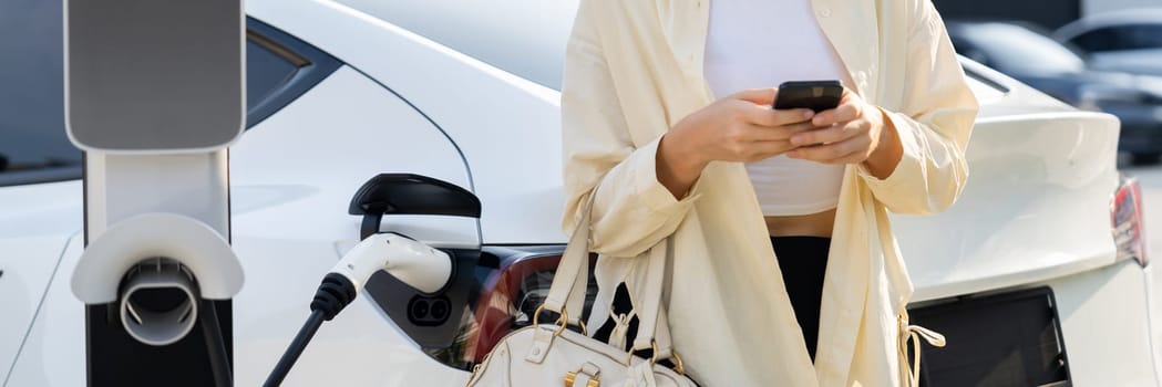 Woman holding shopping bag and use smartphone to pay for electricity to recharge EV car battery from charging station at city mall parking lot. Modern woman go shopping by eco car.Panorama Expedient