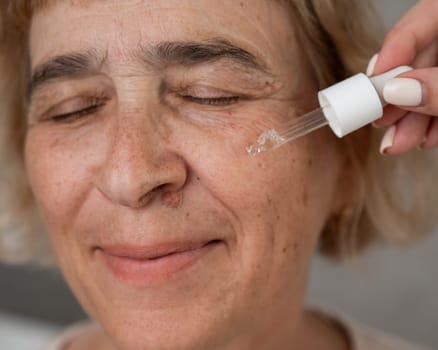 Close-up portrait of an old woman applying hyaluronic acid serum with a pipette. Anti-aging face care