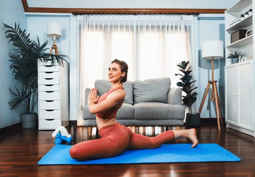 Flexible and dexterity woman in sportswear doing yoga position in meditation posture on exercising mat at home. Healthy gaiety home yoga lifestyle with peaceful mind and serenity.