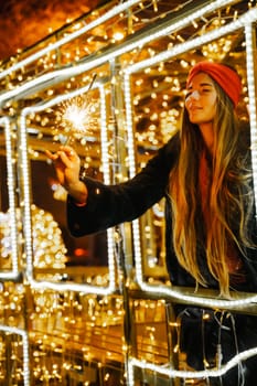 Woman holding sparkler night while celebrating Christmas outside. Dressed in a fur coat and a red headband. Blurred christmas decorations in the background. Selective focus.