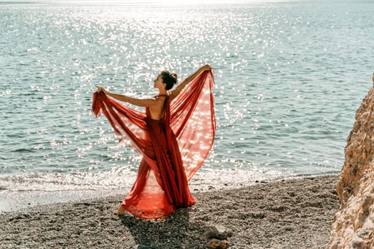 Woman red dress sea. Female dancer in a long red dress posing on a beach with rocks on sunny day. Girl on the nature on blue sky background