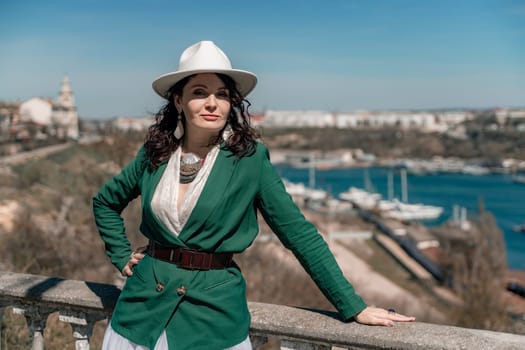 Woman walks around the city, lifestyle. Happy woman in a green jacket, white skirt and hat is sitting on a white fence with balusters overlooking the sea bay and the city