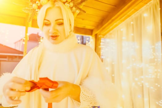 A woman in a white dress is holding a gold box with a red ribbon. She is wearing a crown on her head. The scene takes place in a room with a door and a window. The woman appears to be opening the gift box.