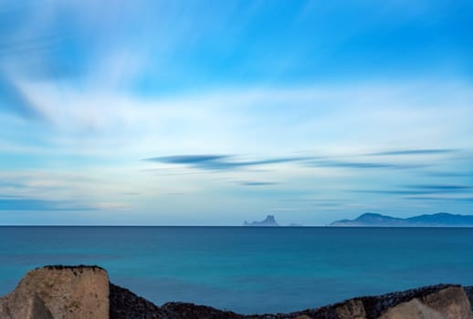 Seascape photo featuring the majestic Es Vedra Island in Ibiza, with smooth water, abstract clouds from long exposure, and solid earth.