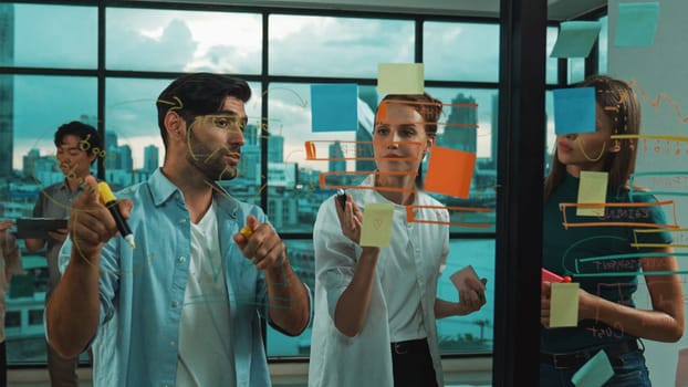 Professional business team brainstorm, sharing, thinking idea while skilled male leader write on sticky notes at glass wall. Skilled manager placed business plan on glass wall at office. Tracery