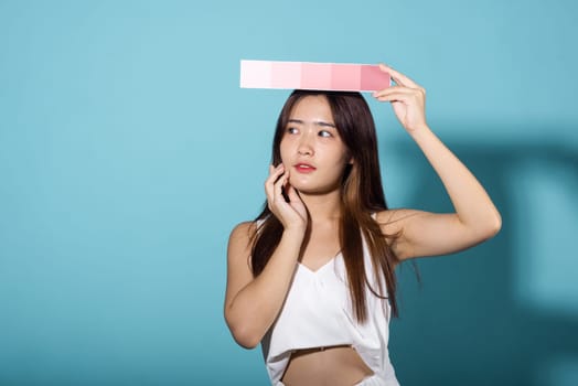 Portrait of happy female hold color card under sun protection, skin beauty face, Asian beautiful young woman smiling and holding skin color scale paper studio shot isolated on blue background