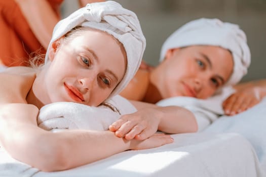 A couple of beautiful young woman with white towel lie on comfortable spa bed during receive relaxing body massage by professional masseur surrounded at spa salon. Gray background. Tranquility.