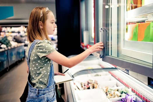 Pretty girl child buying with shopping list in supermarket and looking products in fridge. Beautiful female preteen kid in grocery store