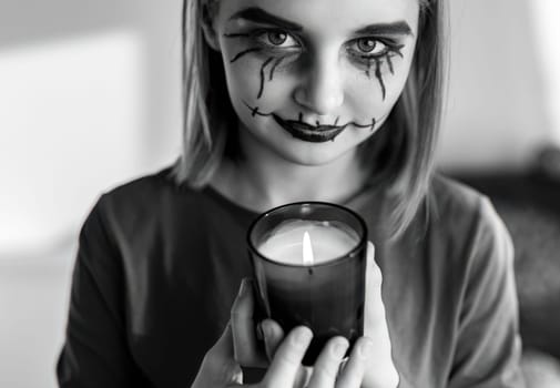 Preteen girl with spooky Halloween makeup posing at camera with lighting candle. Kid's portrait for creepy holiday in black and white.
