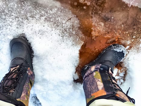 Feet of Hunter or fisherman in big warm boots on winter day on snow. Top view. Fisherman on the ice of a river, lake, reservoir on spring day with melting ice. Dangerous fishing in autumn