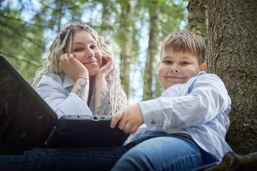 Kirov, Russia - June 12, 2023: Son with laptops in forest And Mom is proud of him. Fat young clever teenage boy with modern IT technologies in nature