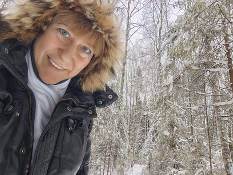 Happy, cheerful middle-aged woman in a warm jacket and a hood with fur taking selfie in winter outdoors. Female Traveler in Alaska, Lapland, Arctic, Siberia