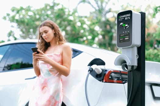 Focused charging station recharging electric vehicle on blurred background of modern woman using smartphone. EV technology utilization for tracking energy usage to optimize battery charging.Synchronos