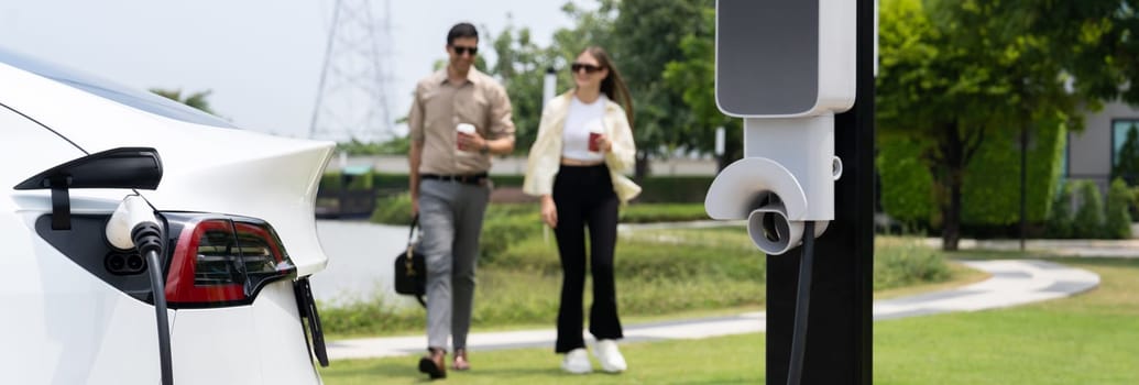 Young couple with coffee recharge EV car battery at charging station connect to electrical industrial power grid. Couple with shopping and travel using eco electric car lifestyle.Panorama Expedient