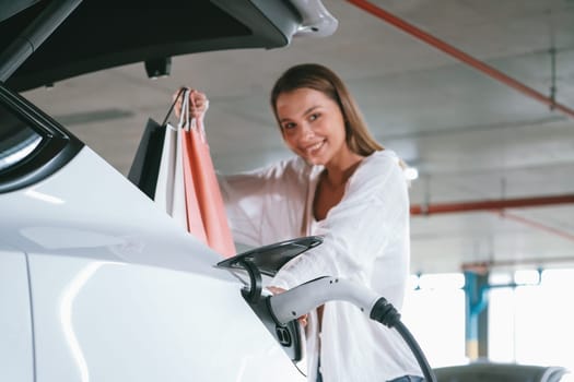 Young woman travel with EV electric car to shopping center parking lot charging in downtown city showing urban sustainability lifestyle by green clean rechargeable energy of electric vehicle innards