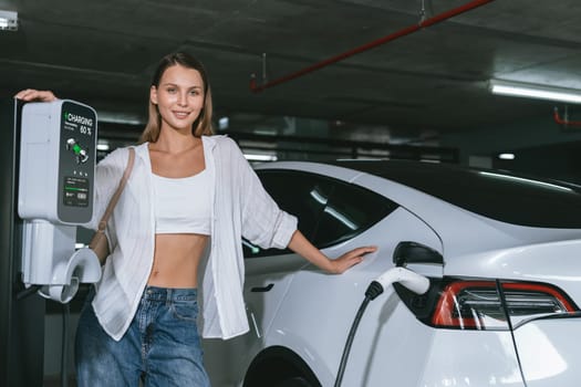 Young woman travel with EV electric car to shopping center parking lot charging in downtown city showing urban sustainability lifestyle by green clean rechargeable energy of electric vehicle innards