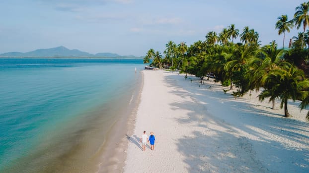Drone view at a couple walking on the white sandy tropical beach of Koh Muk with palm trees soft white sand, and a turqouse colored ocean in Koh Mook Trang Thailand, top view at a couple on the beach