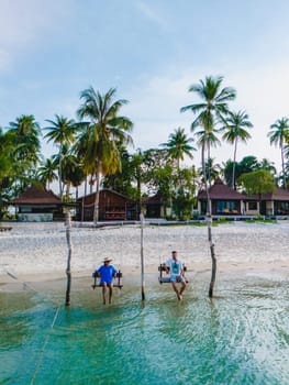 a couple of men and woman on a swing at the beach of Koh Muk a tropical island with palm trees soft white sand, and a turqouse colored ocean in Koh Mook Trang Thailand, couple on a swing in the ocean