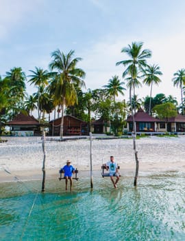 a couple of men and a woman on a swing at the beach of Koh Muk a tropical island with palm trees soft white sand, and a turqouse ocean, Koh Mook Trang Thailand, a couple on a swing in the ocean sunset