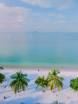 Drone view at a couple walking on the white sandy tropical beach of Koh Muk with palm trees soft white sand, and a turqouse colored ocean in Koh Mook Trang Thailand at summer