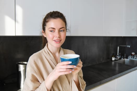 Lifestyle concept. Portrait of happy brunette woman in bathrobe, drinking coffee in the kitchen, having morning cuppa and smiling.