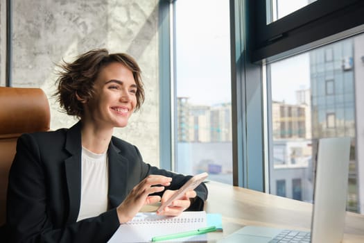 Portrait of businesswoman in suit, sitting in her office in front of window, having a chat on her mobile phone, suing smartphone, has her laptop and documents on table.