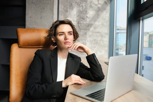 Portrait of confident businesswoman in office, sitting with laptop, looking serious outside window, thinking, solving issue at work.